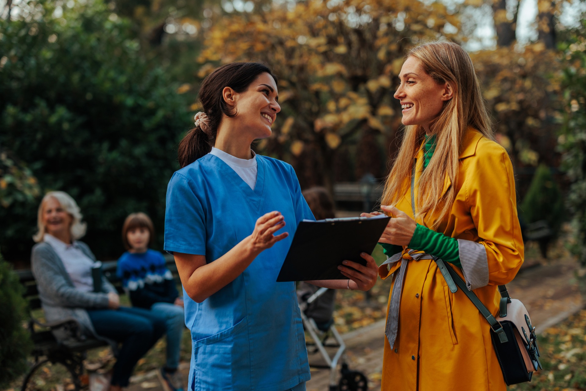Two women talking outdoors, one wearing blue scrubs and holding a clipboard